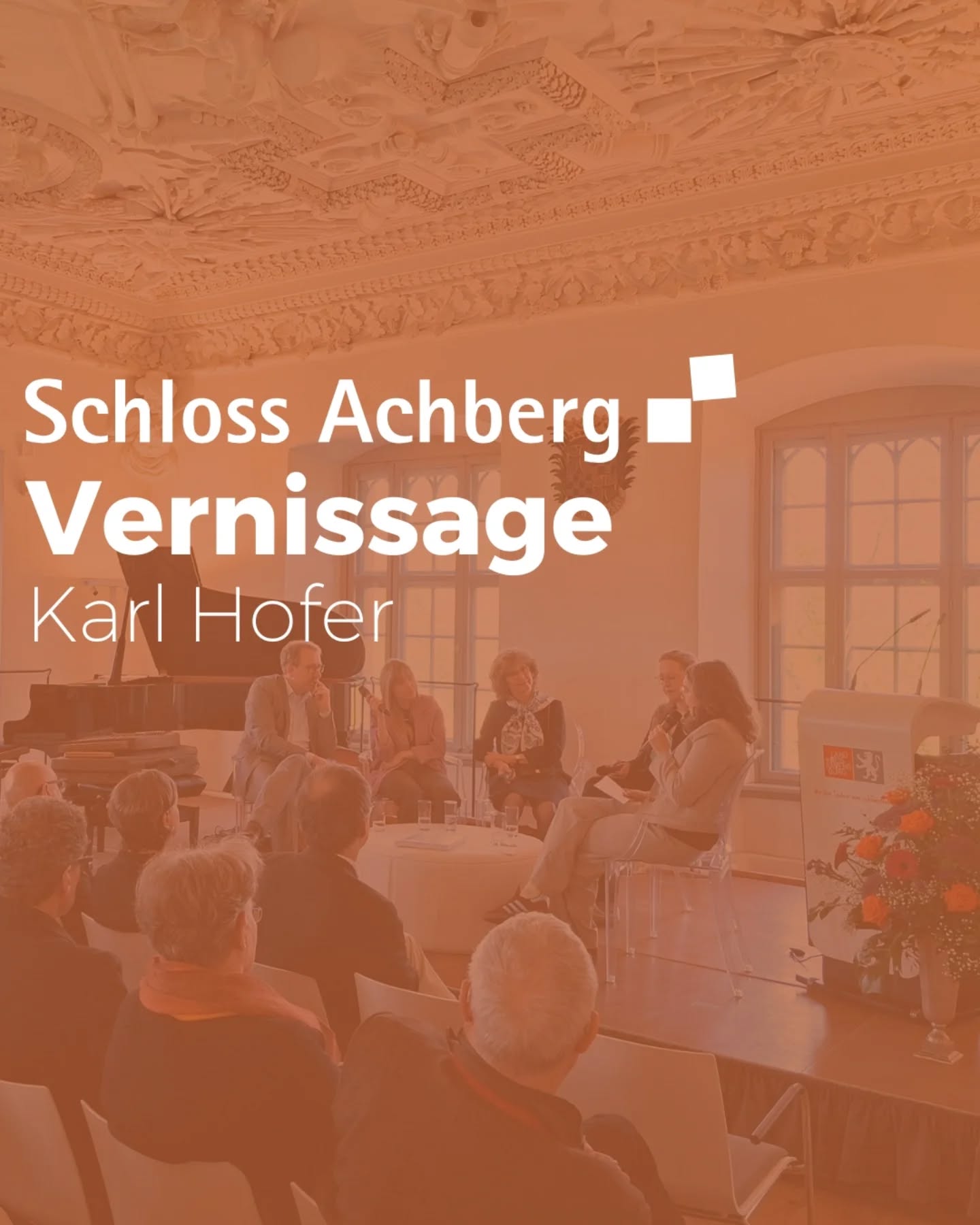 Dieser Alternativtext wurde KI-generiert: Innenansicht eines Raumes mit Stuckdecken und Fenstern. Mehrere Personen sitzen an einem Tisch und scheinen an einer Veranstaltung teilzunehmen, vor der ein Blumenstrauß steht.