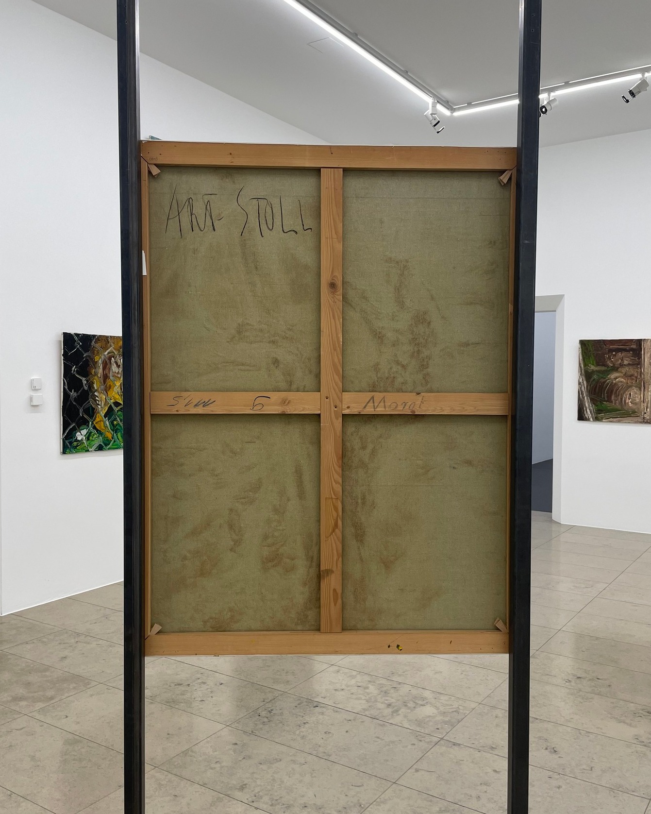 Dieser Alternativtext wurde KI-generiert: Ein Bild hängt mit der Rückseite zum Betrachter. Im Hintergrund sind Teile eines hellen Raumes mit weiteren Bildern zu sehen.