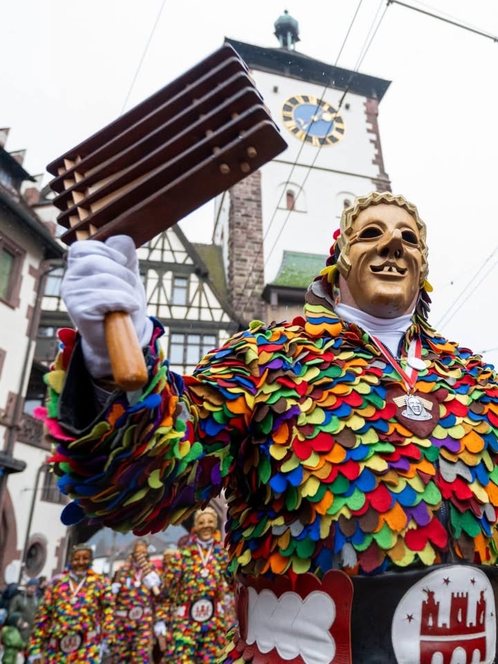 Dieser Alternativtext wurde KI-generiert: Eine Person in einem farbenfrohen Kostüm mit einer Maske hält ein Musikinstrument. Im Hintergrund ist ein Gebäude mit einer Turmuhr zu sehen.