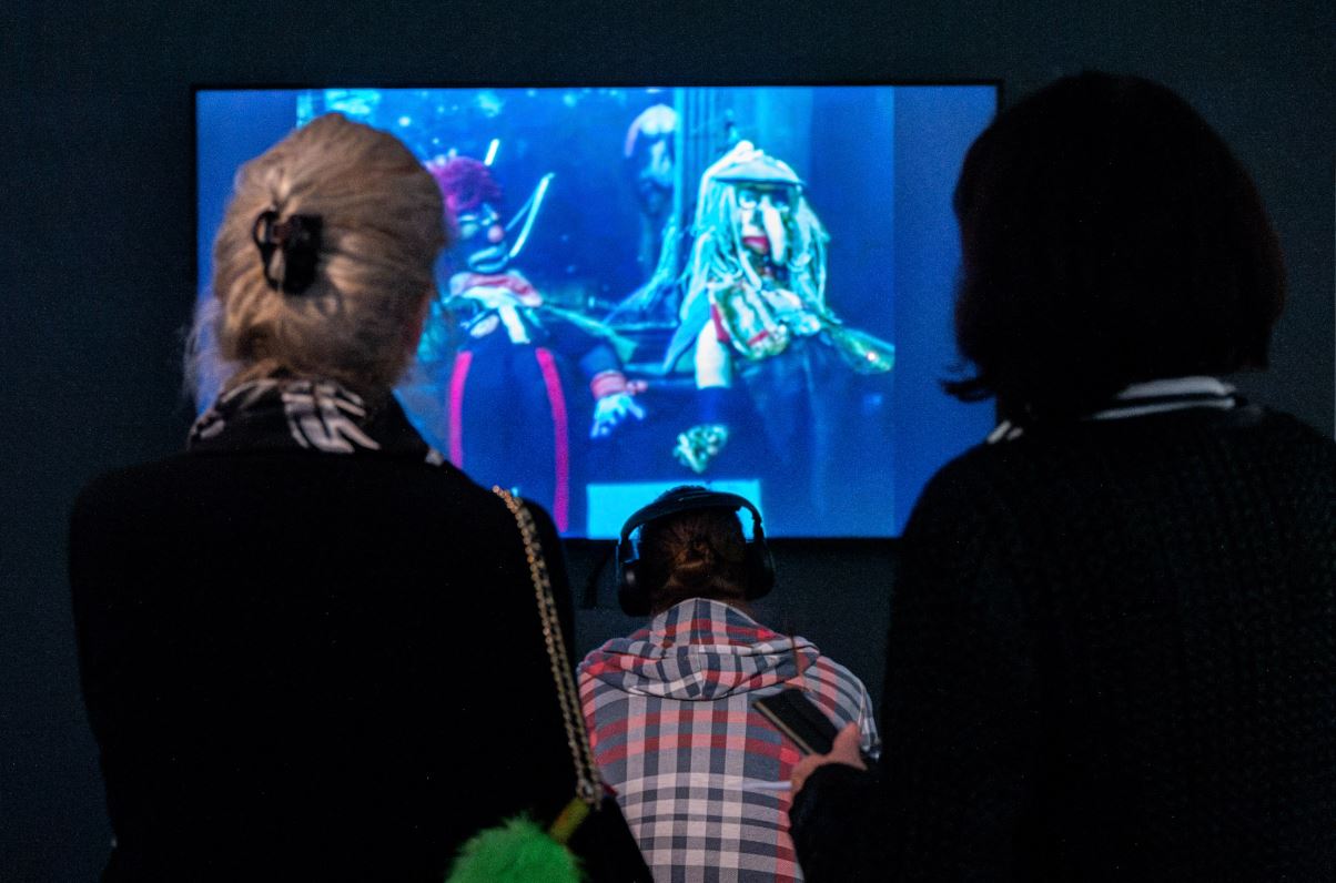 Menschen betrachten einen Bildschirm, auf dem eine Videoinstallation zu sehen ist. Die Szenerie ist dunkel und die Stimmung geheimnisvoll.