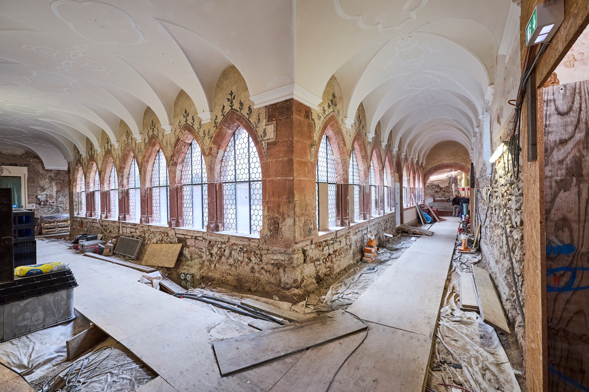 Dieser Alternativtext wurde KI-generiert: Ein langer Raum mit gewölbten Decken und hohen, spitzbogigen Fenstern. Der Raum scheint renoviert oder verlassen zu sein und enthält verschiedene Materialien und Werkzeuge.