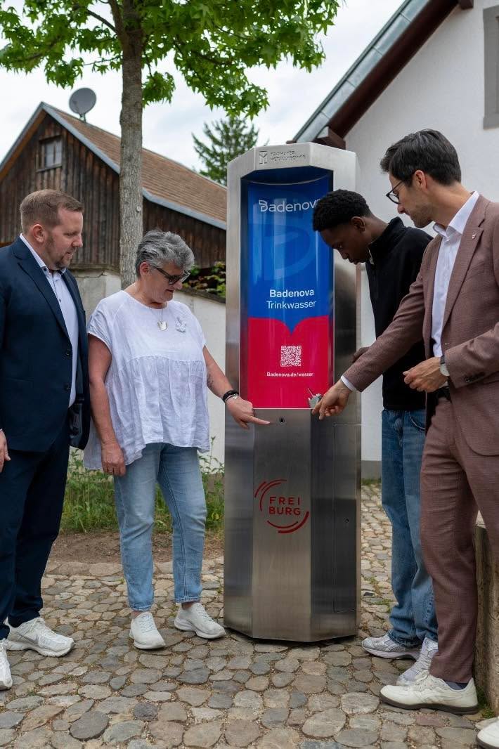 Dieser Alternativtext wurde KI-generiert: Eine Person steht vor einer Wasserspenderanlage mit Werbeanzeigen. Im Hintergrund sind weitere Personen und Gebäude zu sehen.