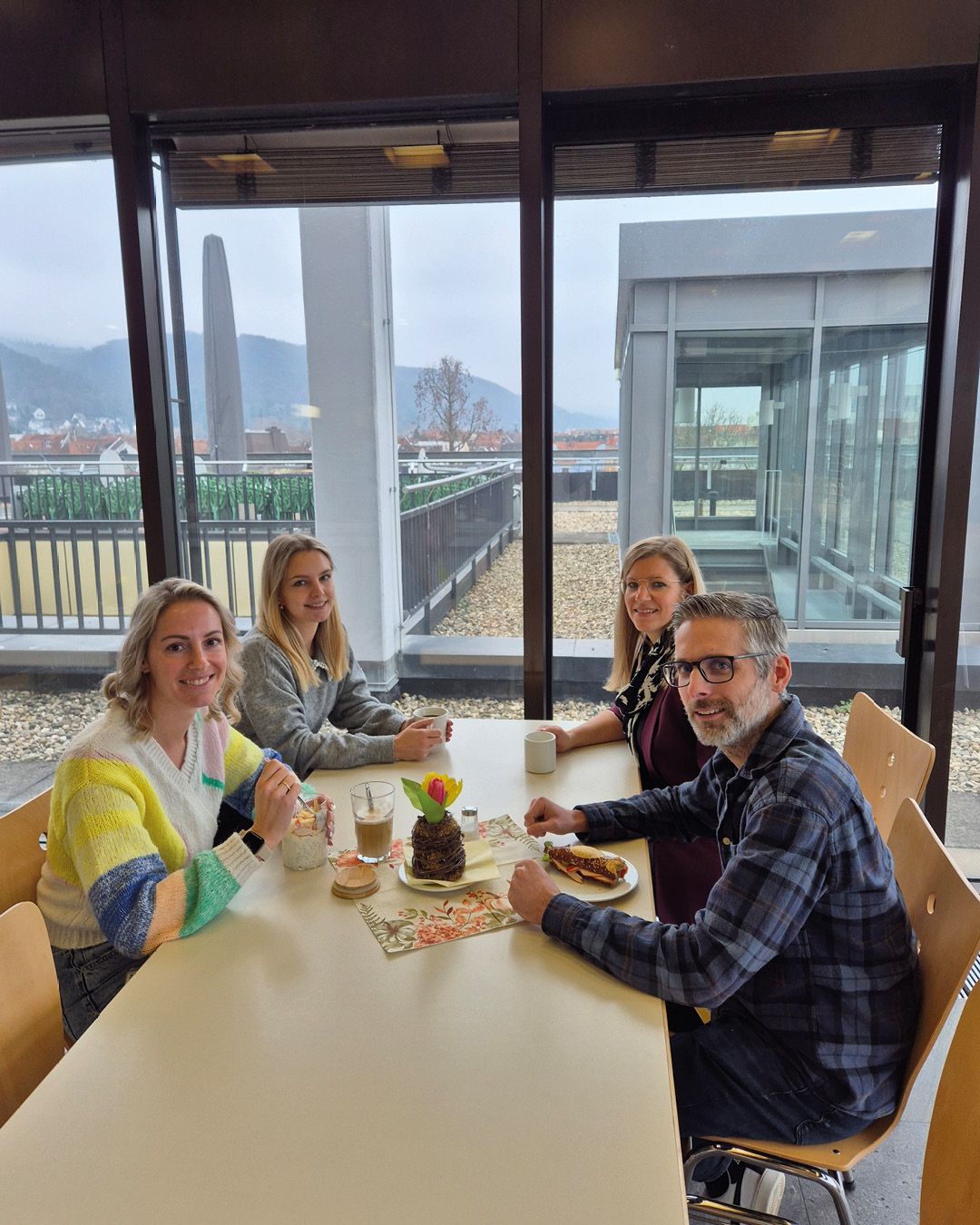 Dieser Alternativtext wurde KI-generiert: Eine Gruppe von Personen sitzt an einem Tisch in der Nähe eines großen Fensters mit Blick auf eine Stadt und Berge. Auf dem Tisch stehen Getränke und Essen.