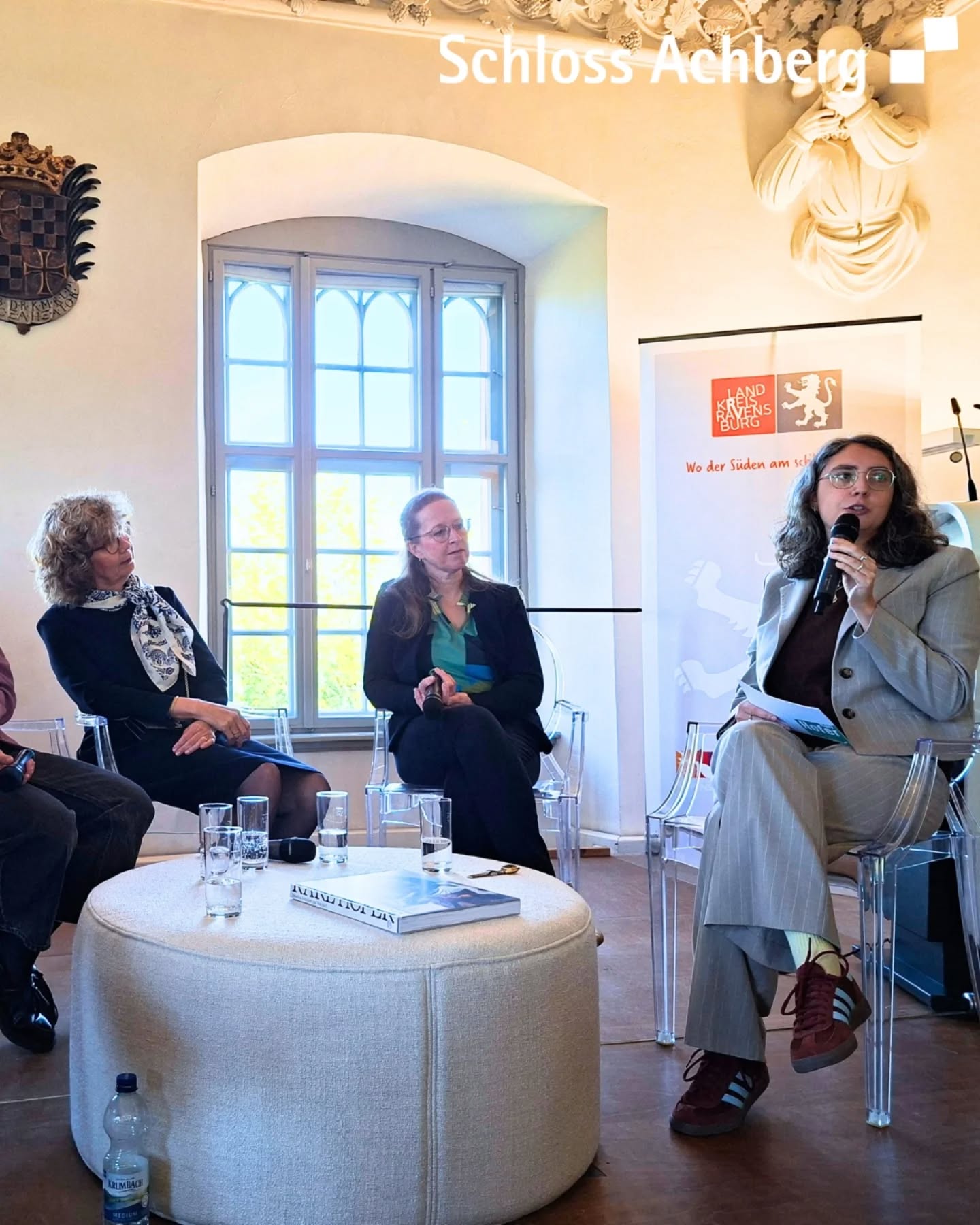 Dieser Alternativtext wurde KI-generiert: Eine Gruppe von Personen sitzt in einem hellen Raum mit einem Fenster im Hintergrund. Ein Tisch mit Wasserflaschen und einem Buch steht im Zentrum.