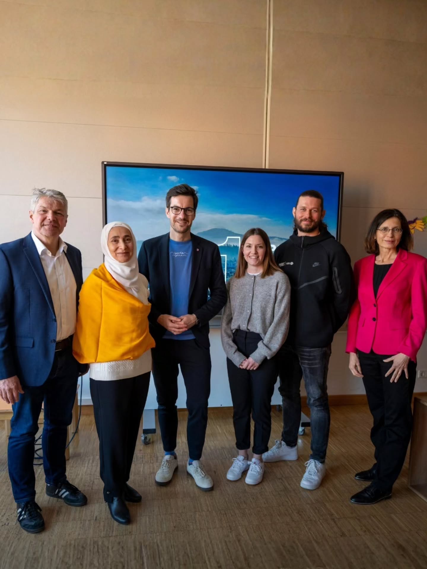 Dieser Alternativtext wurde KI-generiert: Eine Gruppe von sechs Personen steht vor einer Wand und einem großen Bildschirm mit einer Landschaftsdarstellung. Die Aufnahme ist farbig und zeigt Menschen in legerer und formeller Kleidung.