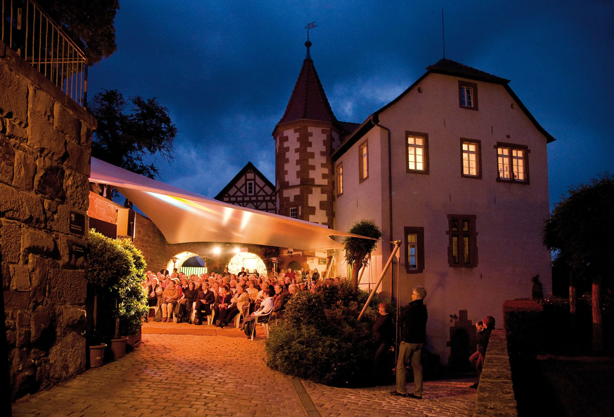 Abendliche Veranstaltung im Innenhof eines historischen Gebäudes mit Turm; Zuschauer sitzen unter einer hell erleuchteten Plane, während im Hintergrund Fenster des Gebäudes leuchten. (KI-Alternativtext)