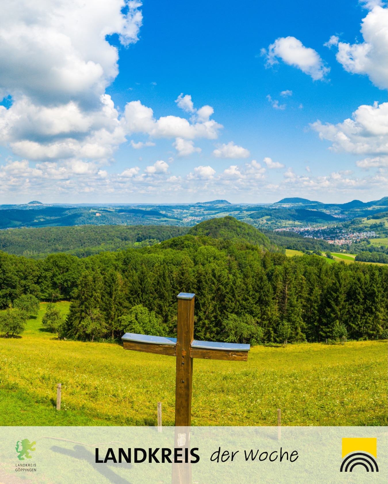 Dieser Alternativtext wurde KI-generiert: Eine farbenfrohe Landschaft mit grünen Wiesen, Bäumen und Hügeln unter einem blauen Himmel mit weißen Wolken. Im Vordergrund steht ein hölzernes Kreuz.