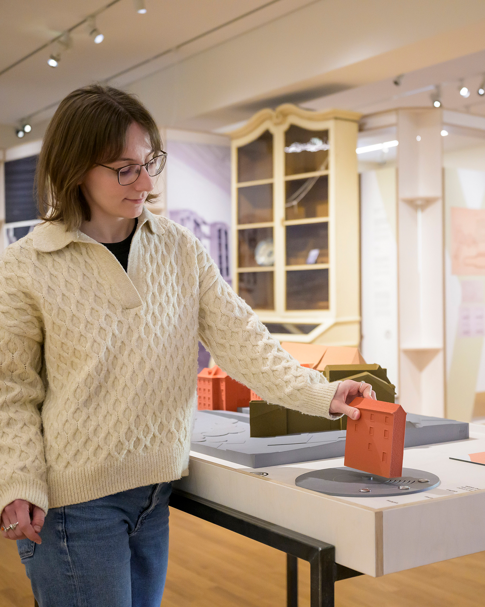 Dieser Alternativtext wurde KI-generiert: Eine Person betrachtet ein Modell eines Gebäudes in einer Ausstellung. Im Hintergrund sind weitere Ausstellungsstücke und Möbel zu sehen.