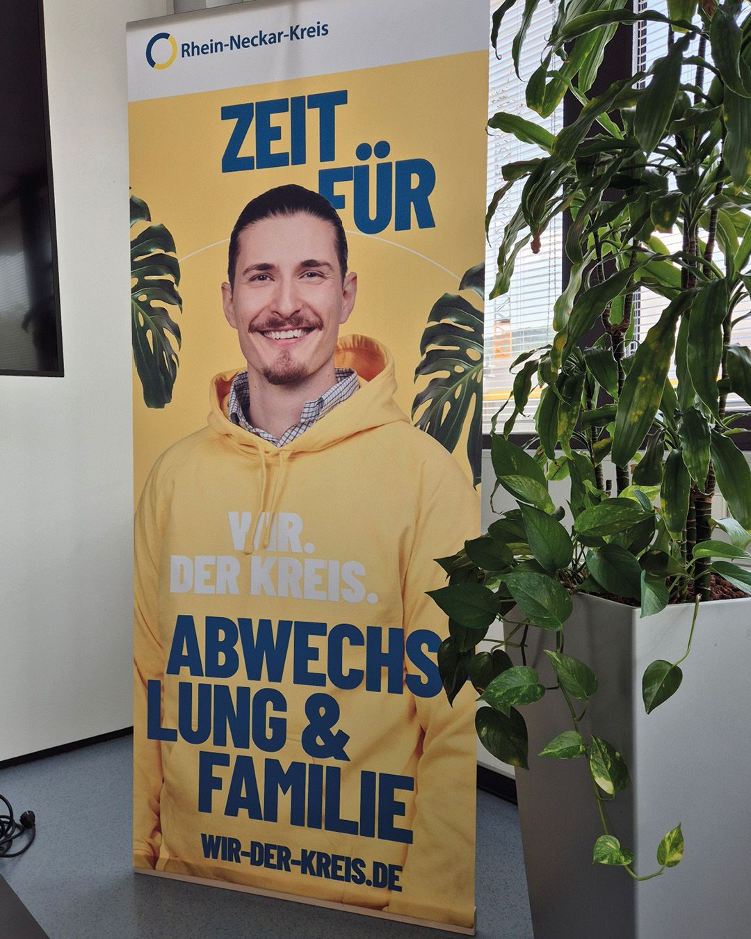 Ein Roll-up-Banner des Rhein-Neckar-Kreises mit gelbem Hintergrund zeigt eine Person in einem gelben Hoodie. Der Text auf dem Banner lautet „ZEIT FÜR“, „WIR. DER KREIS.“, „ABWECHSLUNG & FAMILIE“ und die Webadresse „WIR-DER-KREIS.DE“. Rechts neben dem Banner steht eine große grüne Zimmerpflanze. (KI-Alternativtext)