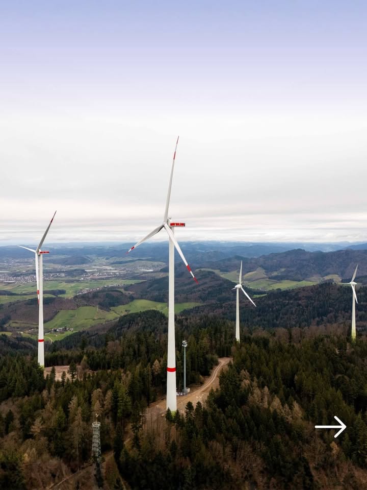 Dieser Alternativtext wurde KI-generiert: Eine Aufnahme aus der Vogelperspektive zeigt mehrere Windräder vor einer hügeligen Landschaft mit Bäumen und einer Stadt in der Ferne. Der Himmel ist bewölkt.