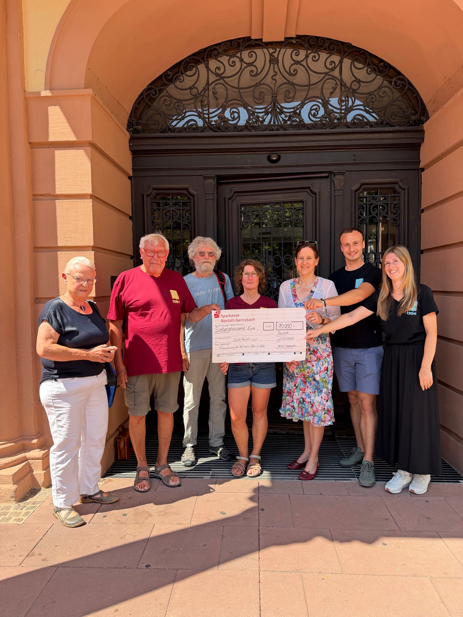 Sieben Personen stehen vor einem großen, dunklen Eingang. Vier Personen in der Mitte halten einen symbolischen Scheck der Sparkasse Rastatt-Gernsbach über 70.000 Euro für die Finanzierung des Festivals tête-à-tête an die Stadt Rastatt. (KI-Alternativtext)