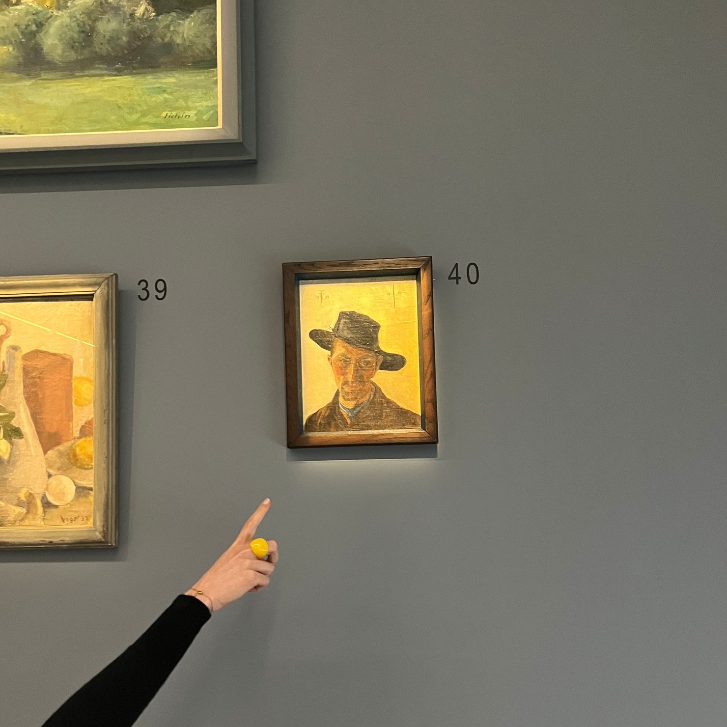 Dieser Alternativtext wurde KI-generiert: Anhand einer hellen Wand hängen mehrere gerahmte Gemälde. Eine Hand mit gelbem Nagellack zeigt auf eines der Bilder.