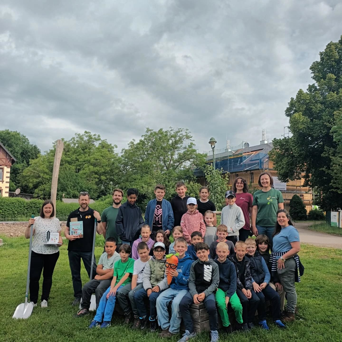 Große Gruppe von Kindern und Erwachsenen im Freien.  Sie halten Werkzeuge und ein Buch.