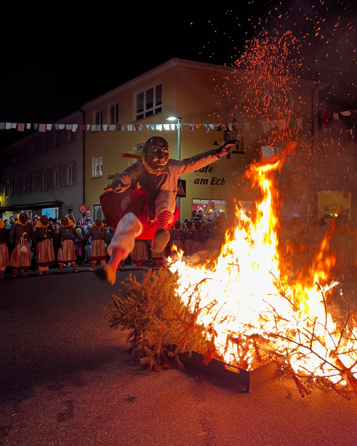 Eine Person springt mit einer Maske und traditioneller Kleidung über ein großes Feuer. Im Hintergrund stehen weitere Personen in Trachten.