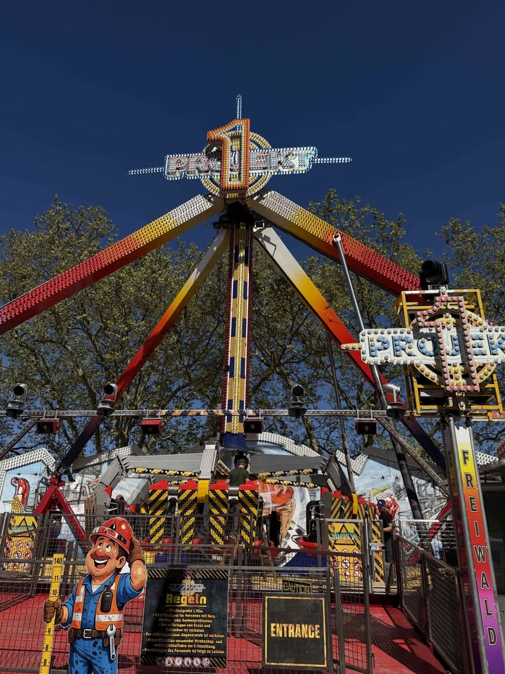 Dieser Alternativtext wurde KI-generiert: Ein hoher Jahrmarkt-Fahrgeschäft mit bunten Lichtern und Dekorationen steht gegen einen blauen Himmel. Im Vordergrund befindet sich eine Figur mit einer Bauarbeiter-Mütze.