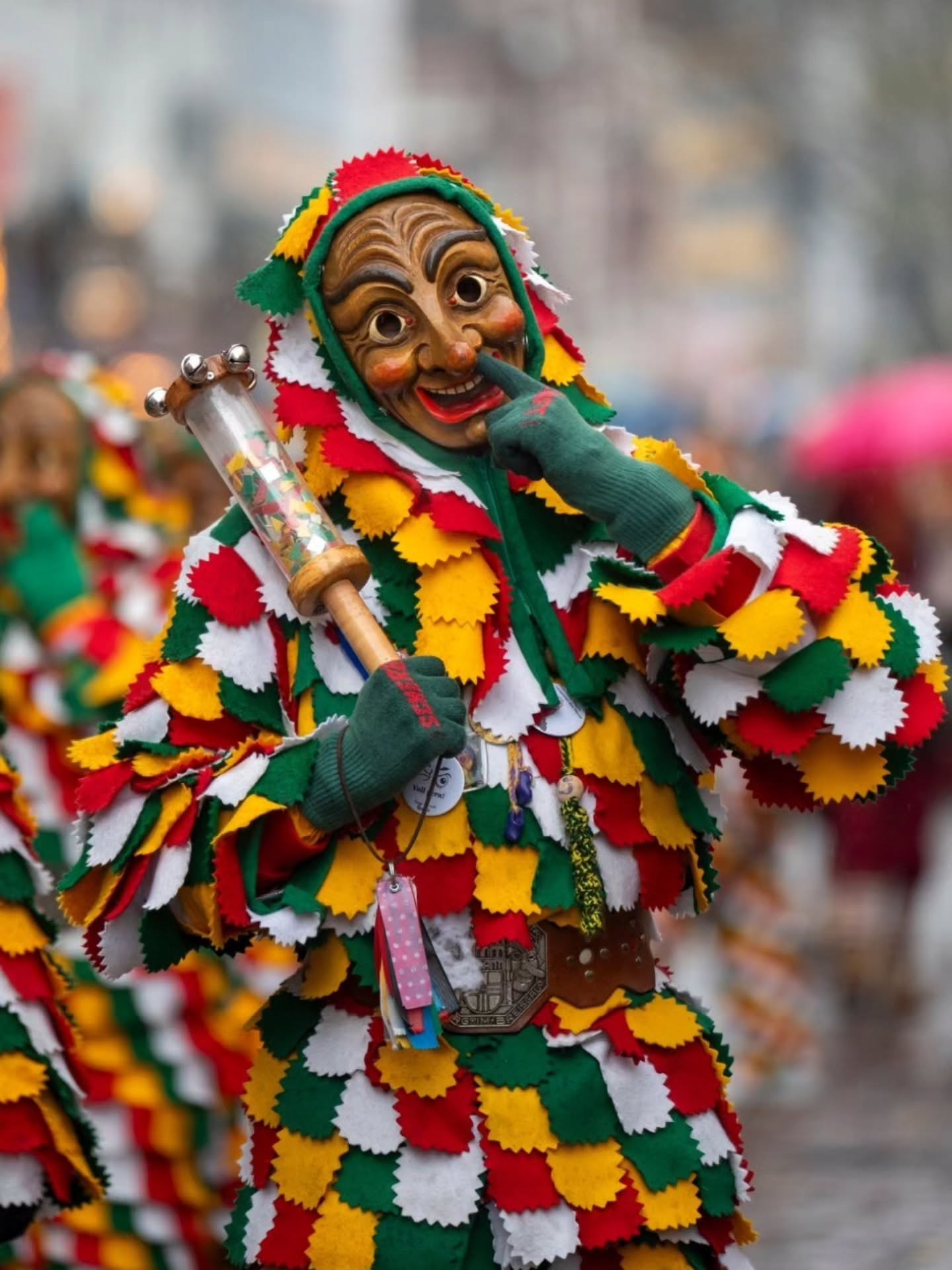 Dieser Alternativtext wurde KI-generiert: Eine Person trägt eine farbenfrohe, traditionelle Maske und ein Kostüm mit Rautenmuster. Im Hintergrund sind weitere verschwommene Gestalten und Farben zu sehen.