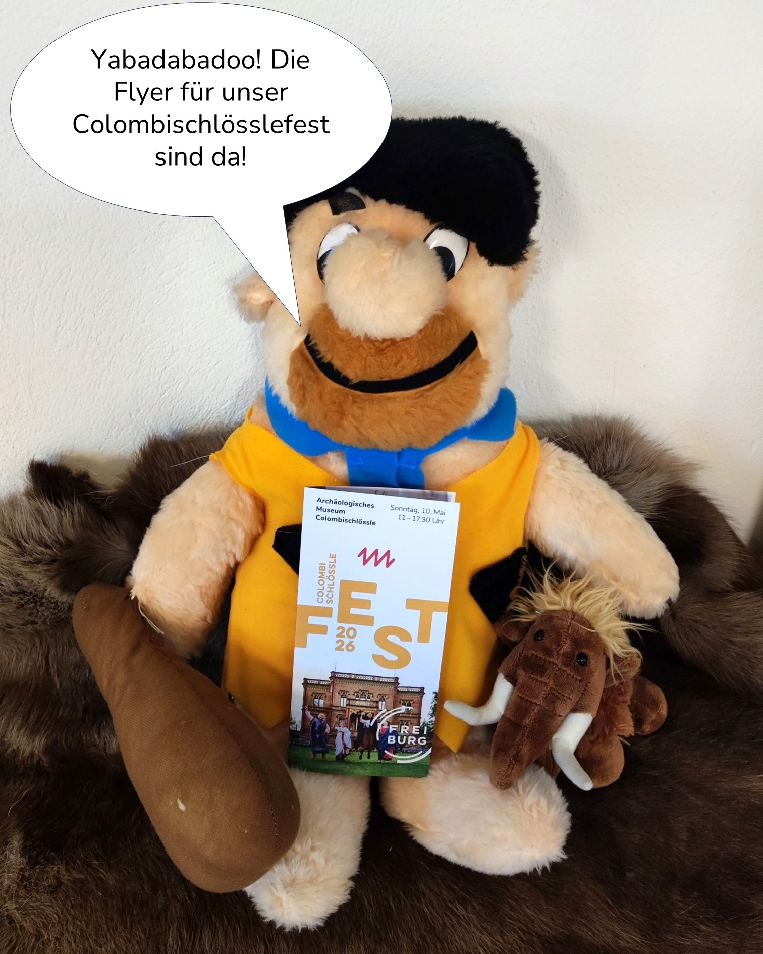 Dieser Alternativtext wurde KI-generiert: Ein Plüschtier in einem Kostüm hält ein Werbeflyer und einen Kalender. Der Flyer bewirbt eine Veranstaltung und zeigt ein Bild einer Burg.