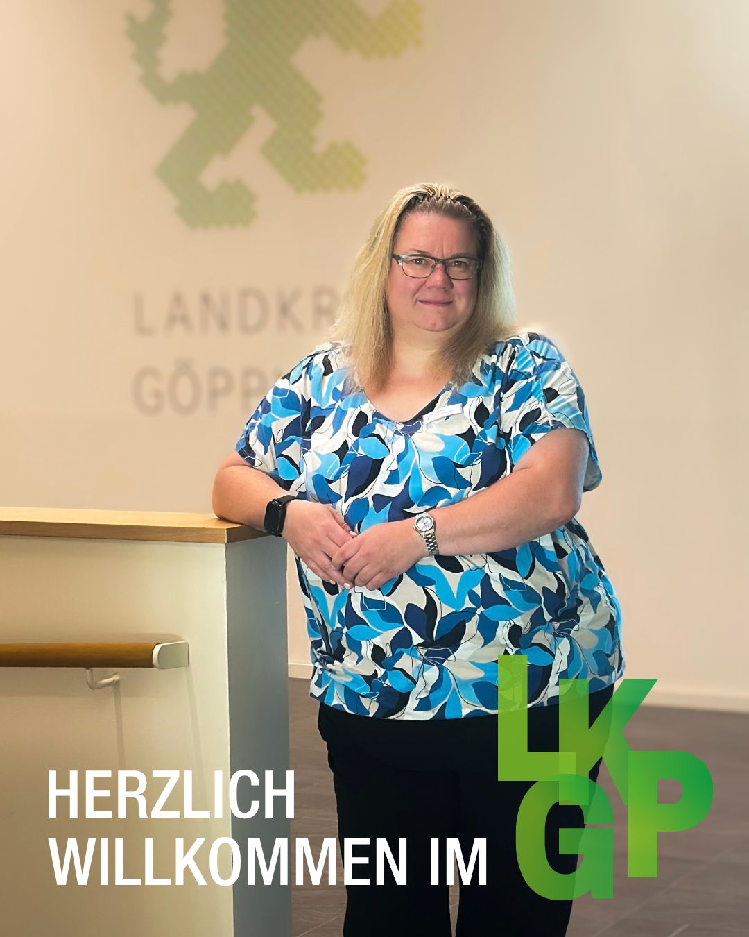 Eine Person mit blonden Haaren und Brille steht vor einer Wand mit einem grünen Logo und dem Text "LANDKR GÖPP". Die Person trägt ein blau-weiß gemustertes Oberteil und schwarze Hosen. Im Vordergrund steht der Text "HERZLICH WILLKOMMEN IM" neben dem großen grünen Logo "LK GP". (KI-Alternativtext)