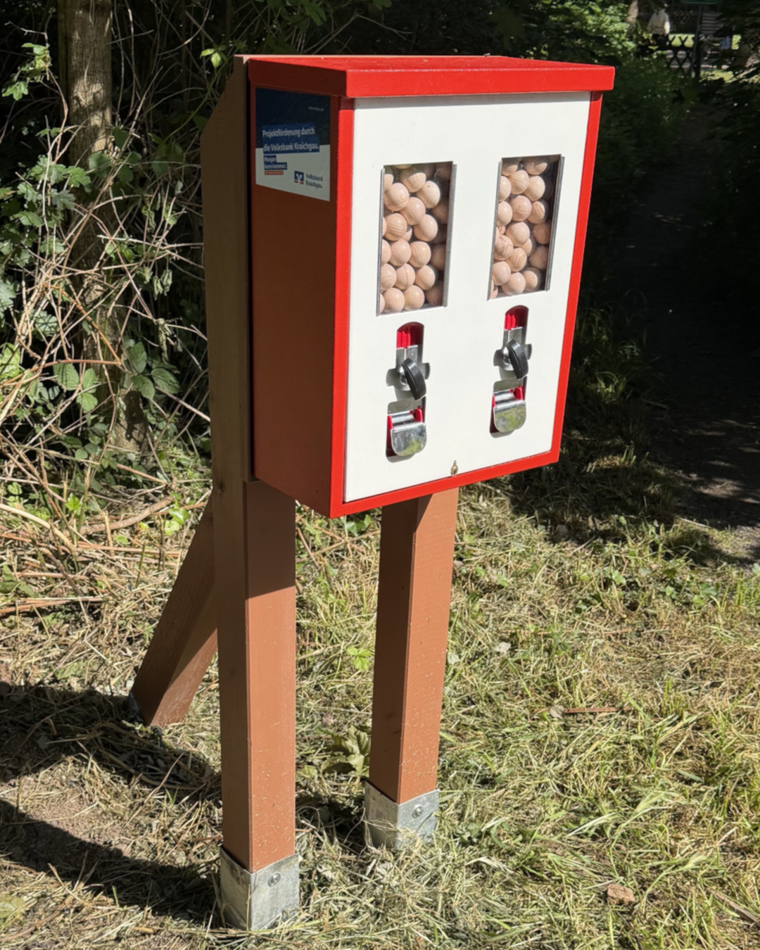 Roter und weißer Holzautomat mit zwei Fächern voller Holzkugeln und Drehhebeln, aufgestellt im Freien auf Gras neben Büschen. An der Seite ein Schild mit der Aufschrift „Projektförderung durch die Volksbank Kraichgau“. (KI-Alternativtext)
