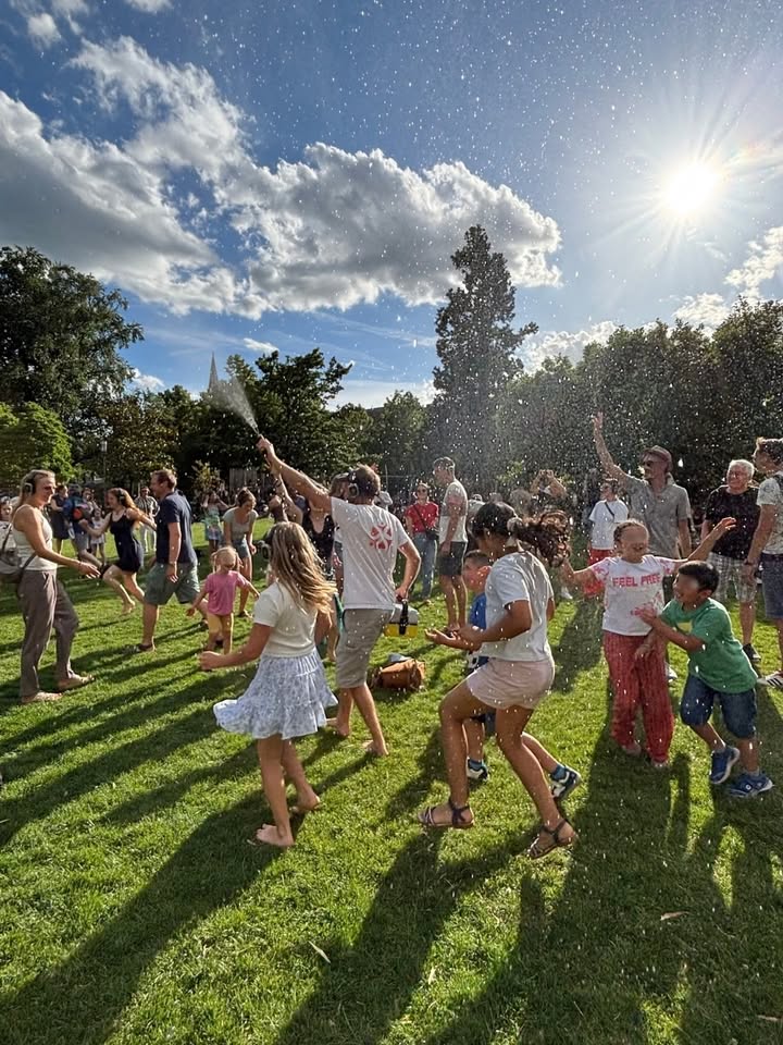 Menschen unterschiedlichen Alters spielen auf einer Wiese im Sonnenschein mit Wasserspritzern, einige halten Sprühflaschen, andere lachen und bewegen sich fröhlich. Im Hintergrund sind Bäume und ein blauer Himmel mit Wolken zu sehen. (KI-Alternativtext)