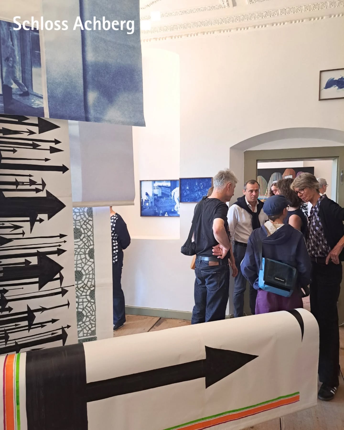 In einem Raum betrachten Personen Kunstwerke. Es gibt Bilder an der Wand und Papierrollen mit Pfeilen und Mustern.