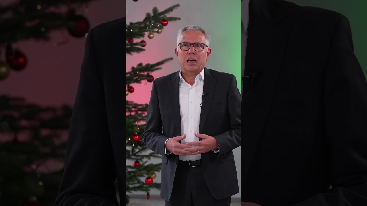Dieser Alternativtext wurde KI-generiert: Eine Person mit Brille in einem dunklen Anzug spricht in die Kamera. Im Hintergrund ist ein festlich geschmückter Weihnachtsbaum zu sehen.