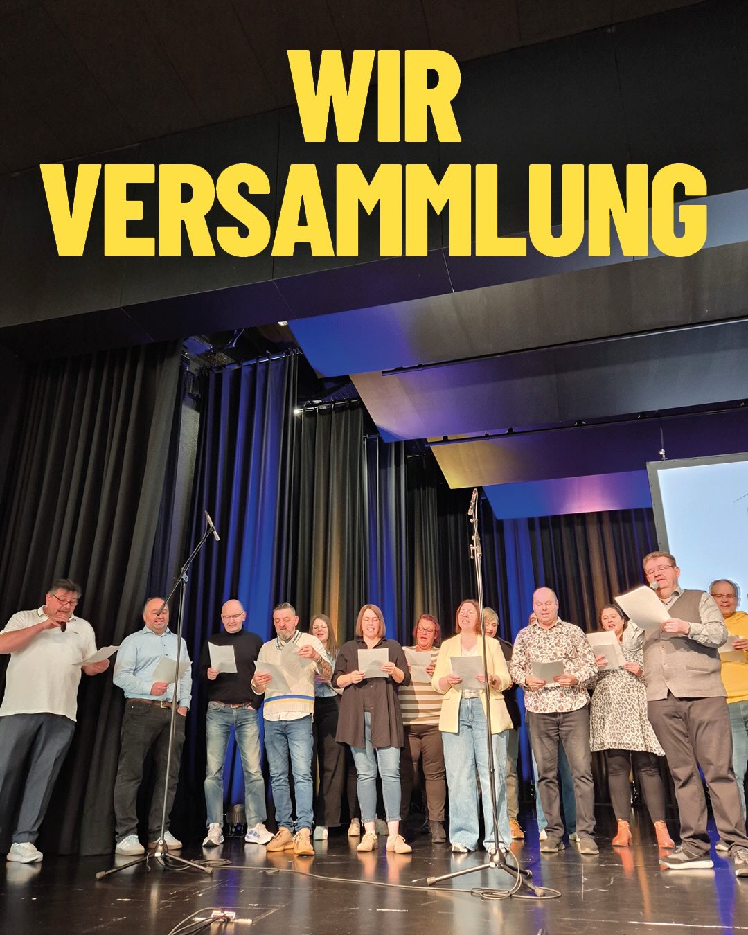 Dieser Alternativtext wurde KI-generiert: Eine Gruppe von Personen steht auf einer Bühne vor blauen Vorhängen und liest von Blättern ab. Über der Bühne ist ein großes Schild mit der Aufschrift 'WIR VERSAMMLUNG' zu sehen.