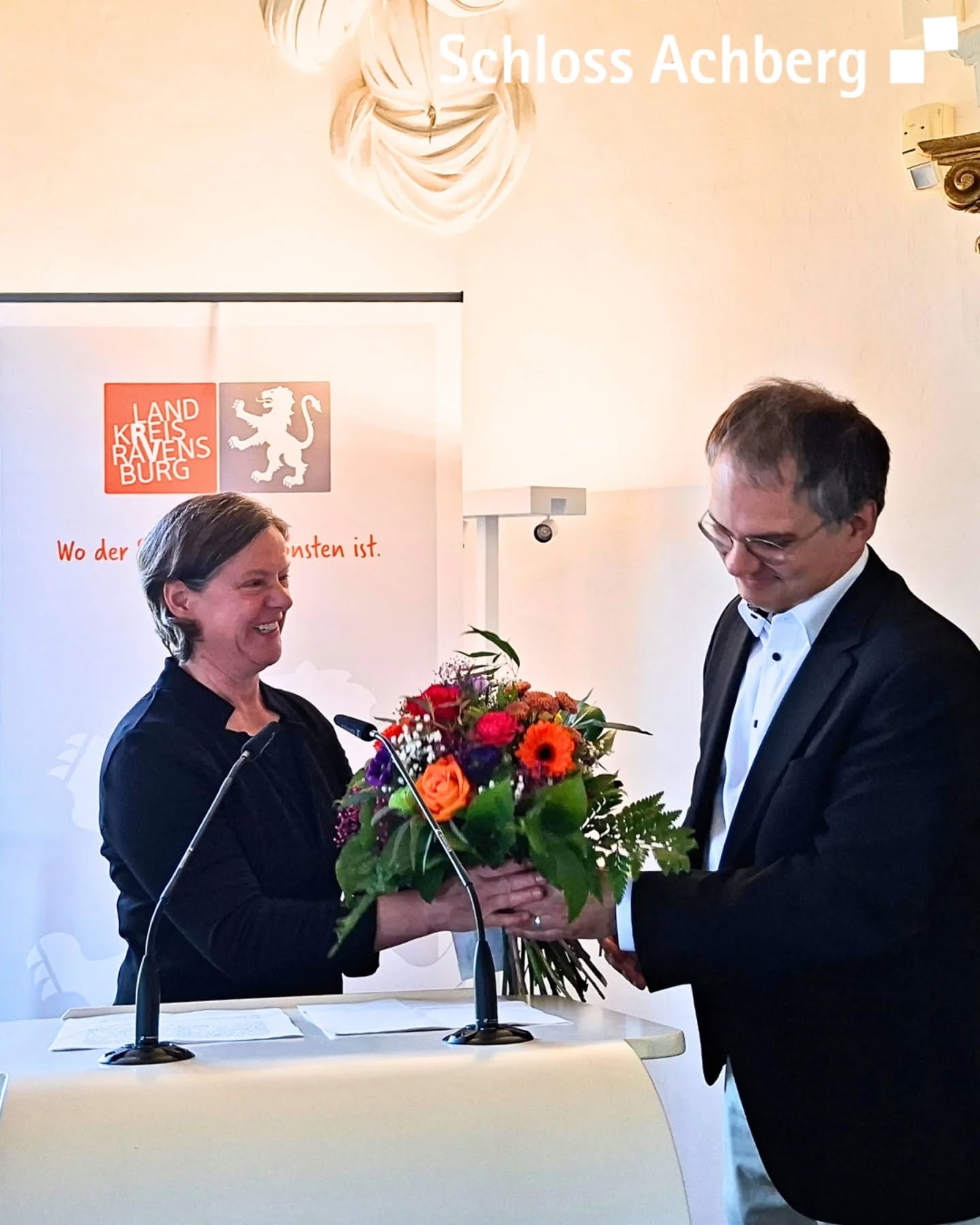 Dieser Alternativtext wurde KI-generiert: Zwei Personen stehen vor einem Tisch mit Blumenschmuck. Im Hintergrund sind Wappen und eine Flagge zu sehen.