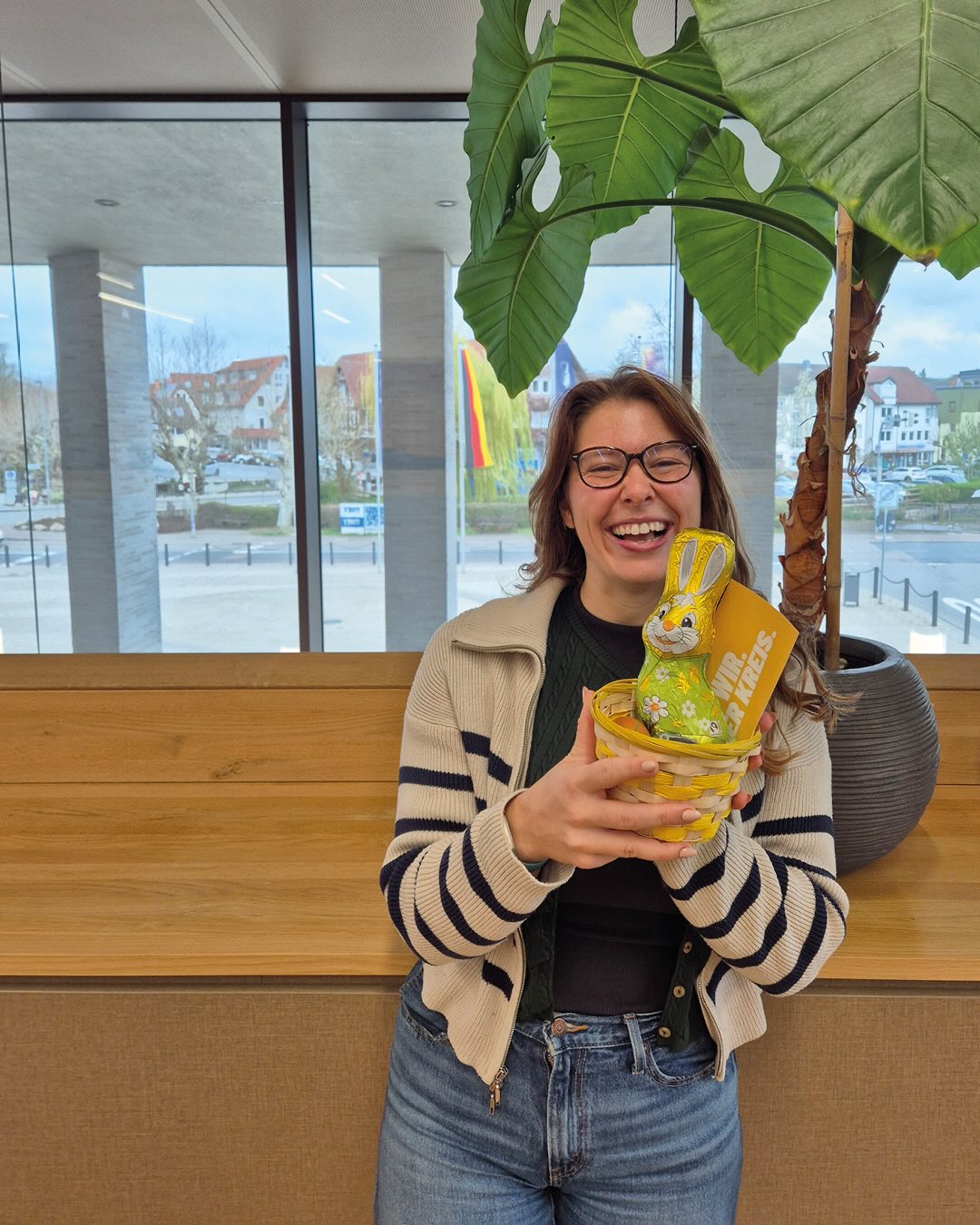 Dieser Alternativtext wurde KI-generiert: Eine Person steht lächelnd in einem hellen Raum mit einer Pflanze im Hintergrund. Sie hält eine bunt verzierte, osterthematische Überraschungseierschachtel in den Händen.