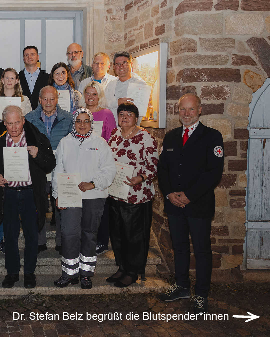 Dieser Alternativtext wurde KI-generiert: Eine Gruppe von Menschen steht auf Stufen vor einer steinernen Wand. Einige halten Urkunden in den Händen, andere tragen Abzeichen oder Uniformen.