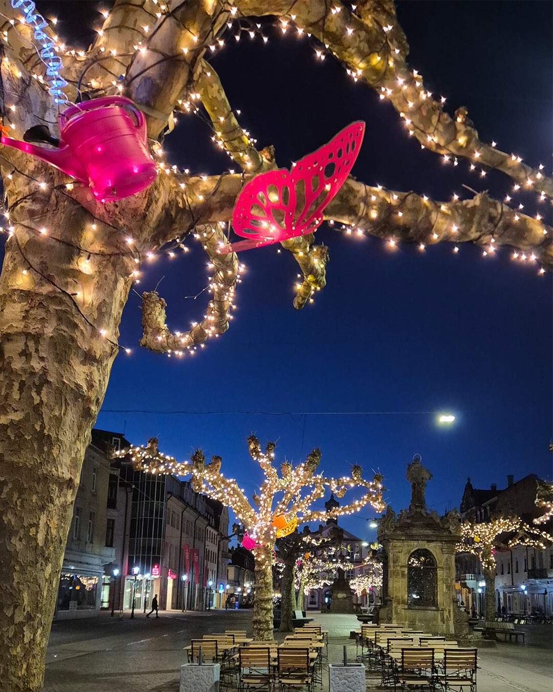 Dieser Alternativtext wurde KI-generiert: Ein Baum ist mit Lichtern und bunten Dekorationen in Schmetterlingsform geschmückt. Im Hintergrund ist eine Stadtansicht bei Nacht zu sehen.