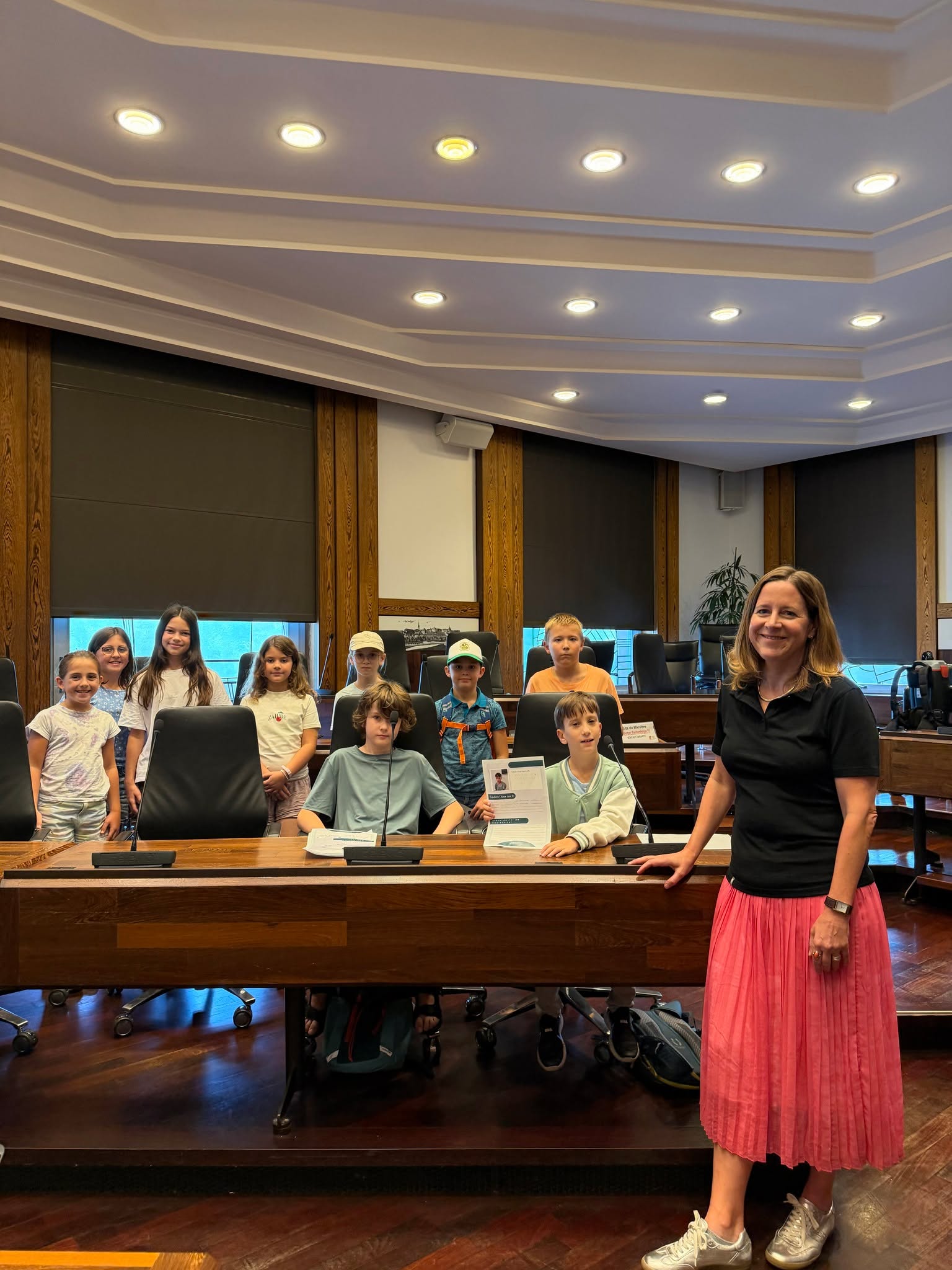 Kinder und eine erwachsene Person posieren in einem Saal mit Holzvertäfelung und Mikrofonen. Einige Kinder sitzen an einem großen Tisch, während andere dahinter stehen. Ein Kind hält ein Dokument mit der Aufschrift "TalentCamp 2023" in die Höhe. (KI-Alternativtext)