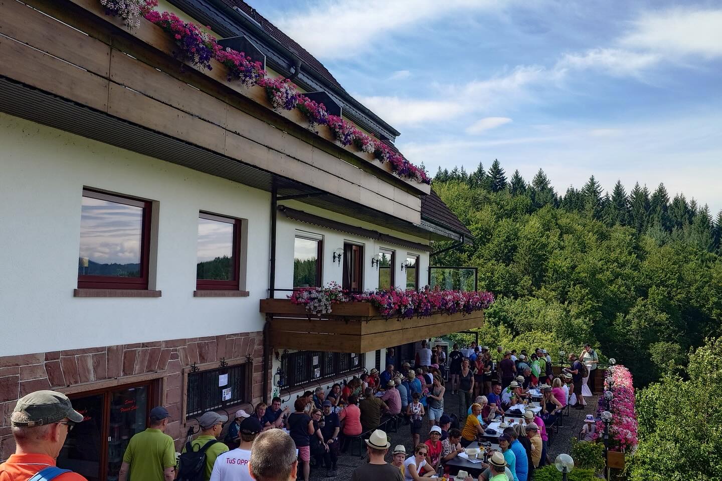 Eine große Gruppe von Personen versammelt sich vor einem Gebäude mit blumengeschmückten Balkonen und Fenstern, mit einem bewaldeten Hintergrund unter einem bewölkten Himmel.