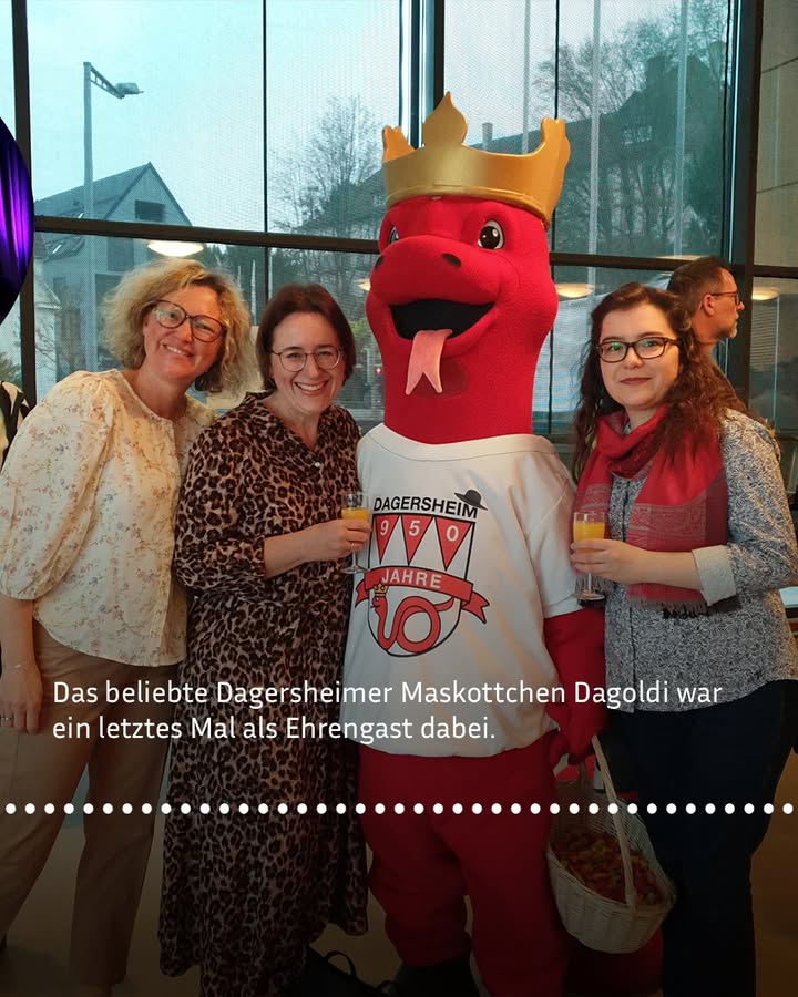Dieser Alternativtext wurde KI-generiert: Drei Personen stehen neben einer rot gekleideten Figur mit einem Wappen. Im Hintergrund ist durch ein Fenster eine Straße zu sehen.