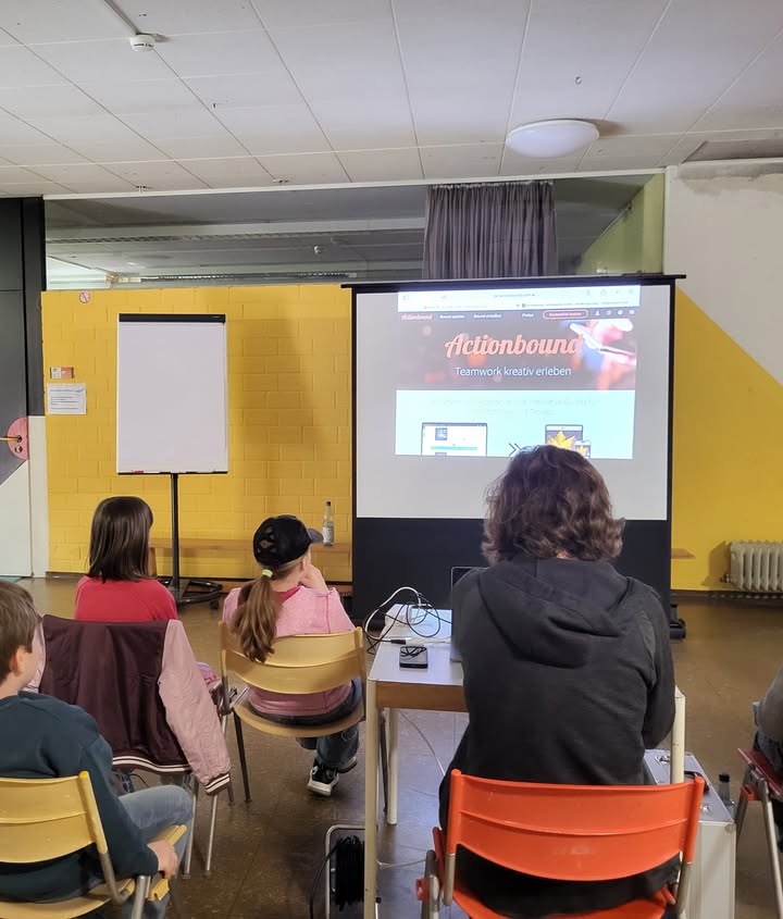 Dieser Alternativtext wurde KI-generiert: Eine Gruppe von Personen sitzt auf Stühlen und schaut auf eine Präsentation an einem Bildschirm. Die Präsentation wird in einem Raum mit gelben Wänden gezeigt.