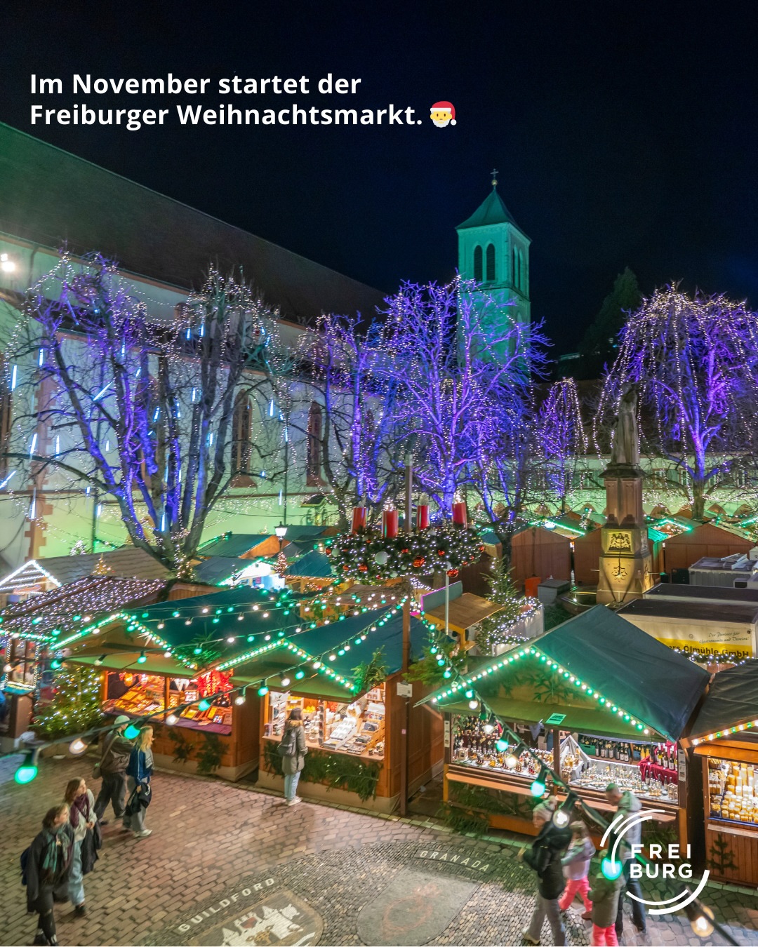 Dieser Alternativtext wurde KI-generiert: Ein nächtliches Bild zeigt festlich erleuchtete Marktstände vor einer Kirche. Über den Ständen und Bäumen erstrecken sich Lichterketten.