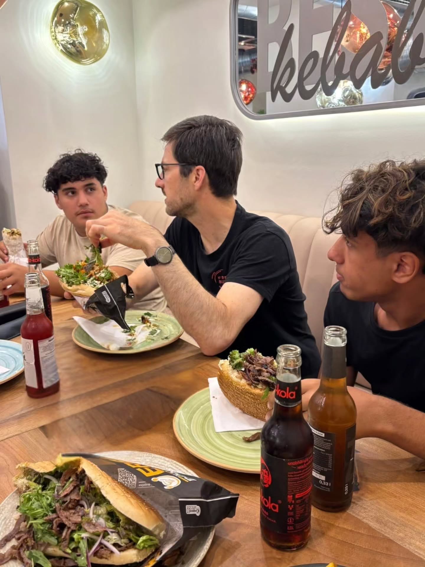 Drei Personen sitzen an einem Holztisch in einem Restaurant und essen große Sandwiches mit Fleisch und Salat. Die Person in der Mitte, die eine Brille und ein schwarzes T-Shirt trägt, hält ein Sandwich in der Hand. Auf dem Tisch stehen mehrere Glasflaschen, darunter eine mit dem Etikett "kola", und im Hintergrund ist ein Schild mit der Aufschrift "kebab" zu sehen. (KI-Alternativtext)