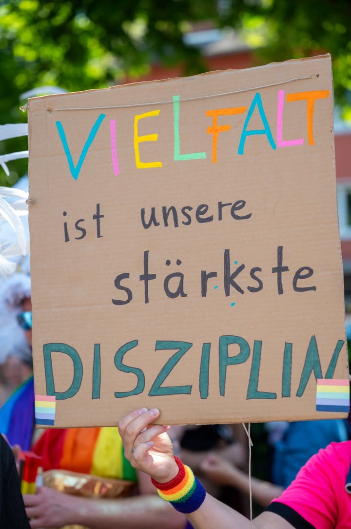 Eine Hand hält ein selbstgemachtes Schild aus Karton mit der Aufschrift „VIELFALT ist unsere stärkste DISZIPLIN“ in bunten Buchstaben, unten rechts und links je eine kleine Regenbogenflagge. Im Hintergrund sind unscharf weitere Personen und grüne Bäume zu erkennen. (KI-Alternativtext)