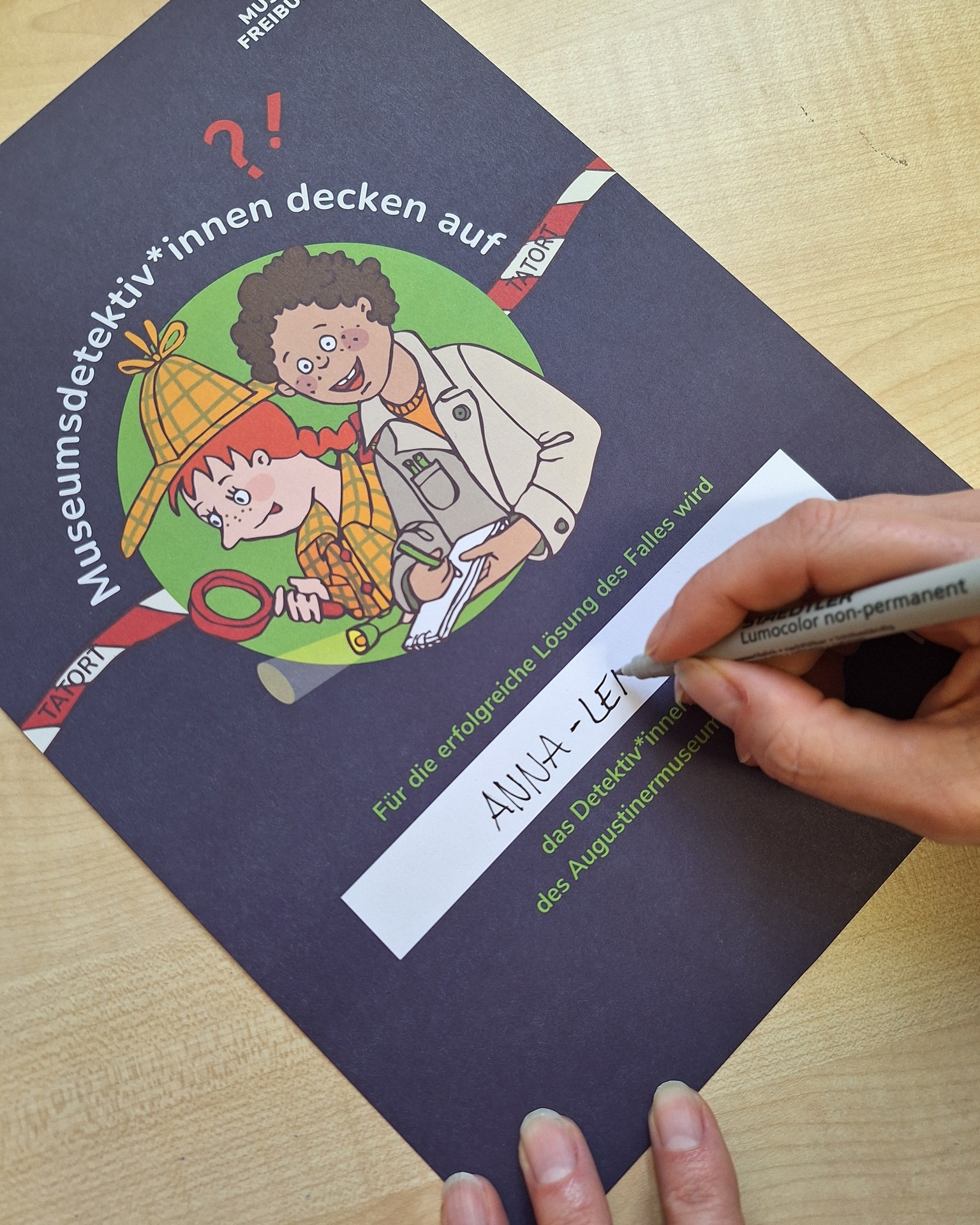 Dieser Alternativtext wurde KI-generiert: Ein Buch mit einer Illustration einer Person mit Hut und Lupe liegt auf einem Tisch. Eine Hand hält einen Stift über eine geöffnete Seite des Buches.