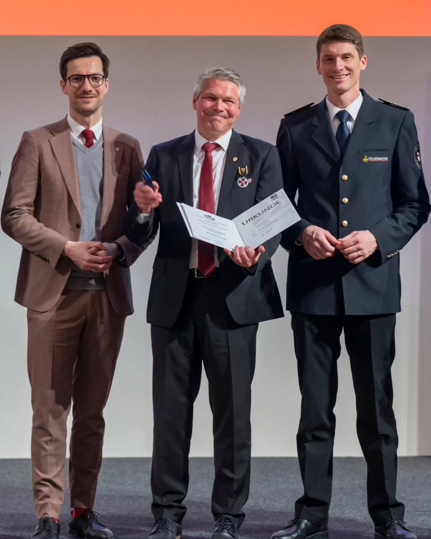 Dieser Alternativtext wurde KI-generiert: Drei Personen stehen nebeneinander auf einer Bühne. Eine Person hält ein Dokument mit der Aufschrift 'URKUNDE' in den Händen.