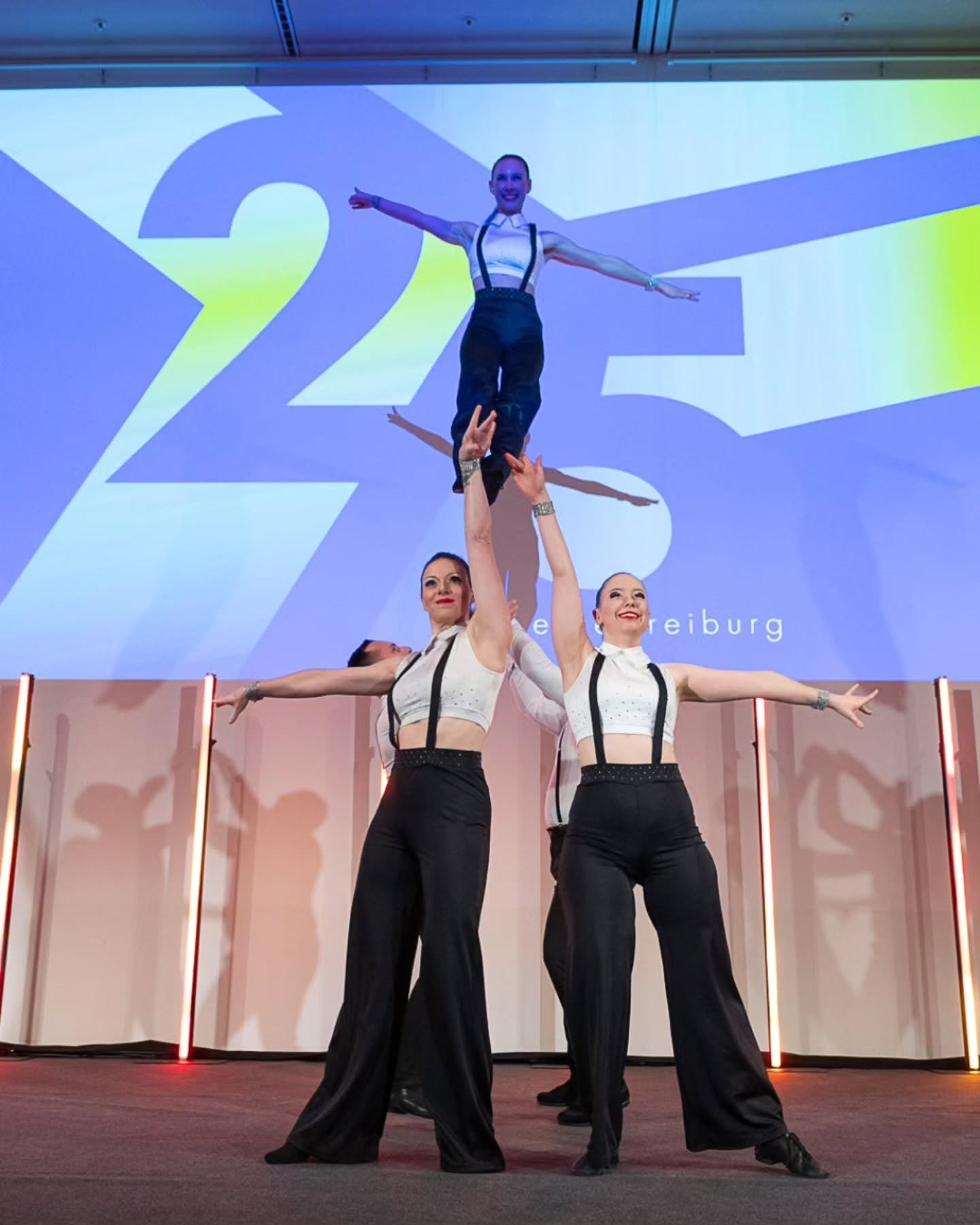 Dieser Alternativtext wurde KI-generiert: Eine Person wird von zwei weiteren Personen gestützt und in der Luft gehalten. Im Hintergrund ist eine große, farbenfrohe Grafik zu sehen.