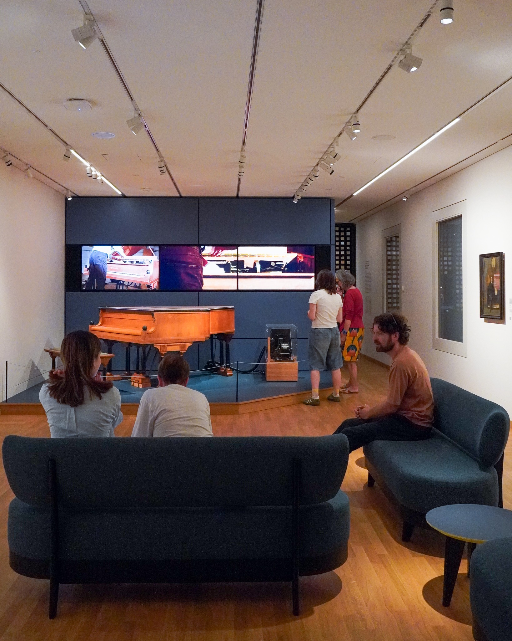 In einem hellen Ausstellungsraum sitzen drei Personen auf dunklen Sofas im Vordergrund und blicken auf eine erhöhte Plattform mit einem historischen Klavier. Dahinter zeigt ein großer Bildschirm ein Video von spielenden Händen, vor dem zwei weitere Personen stehen. Rechts an der Wand hängt ein gerahmtes Gemälde. (KI-Alternativtext)