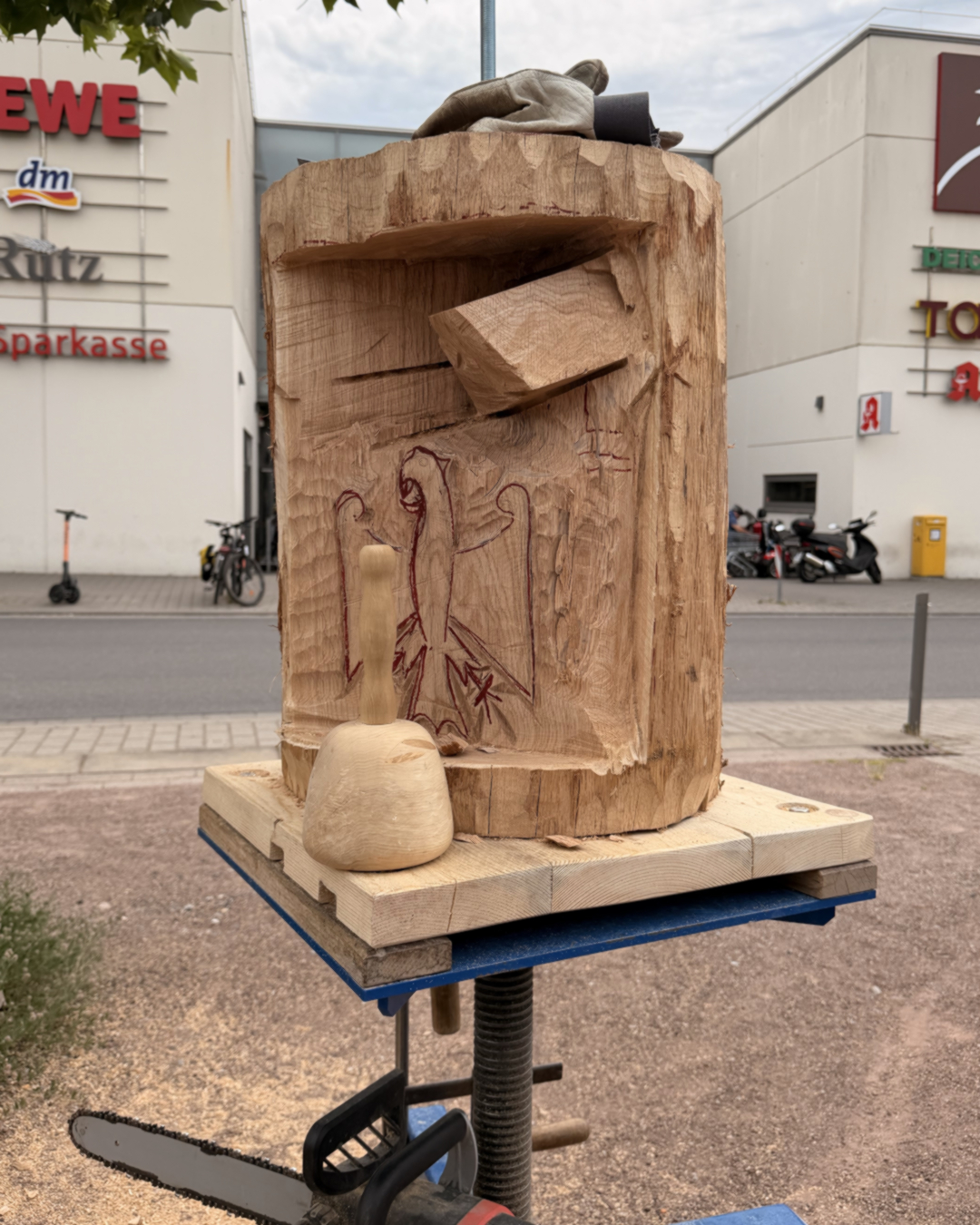 Ein großer Holzstamm auf einer hölzernen Plattform, teilweise geschnitzt mit roten Linien und einem herausgearbeiteten Block, daneben ein Holzhammer; im Hintergrund Geschäfte und eine Kettensäge. (KI-Alternativtext)