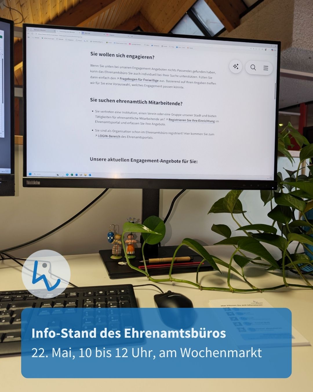 Computerbildschirm mit einer Webseite zum Ehrenamt, darunter ein blauer Hinweis auf einen Info-Stand des Ehrenamtsbüros am 22. Mai von 10 bis 12 Uhr am Wochenmarkt. Auf dem Schreibtisch daneben eine Pflanze, kleine Figuren und ein Keyboard. (KI-Alternativtext)