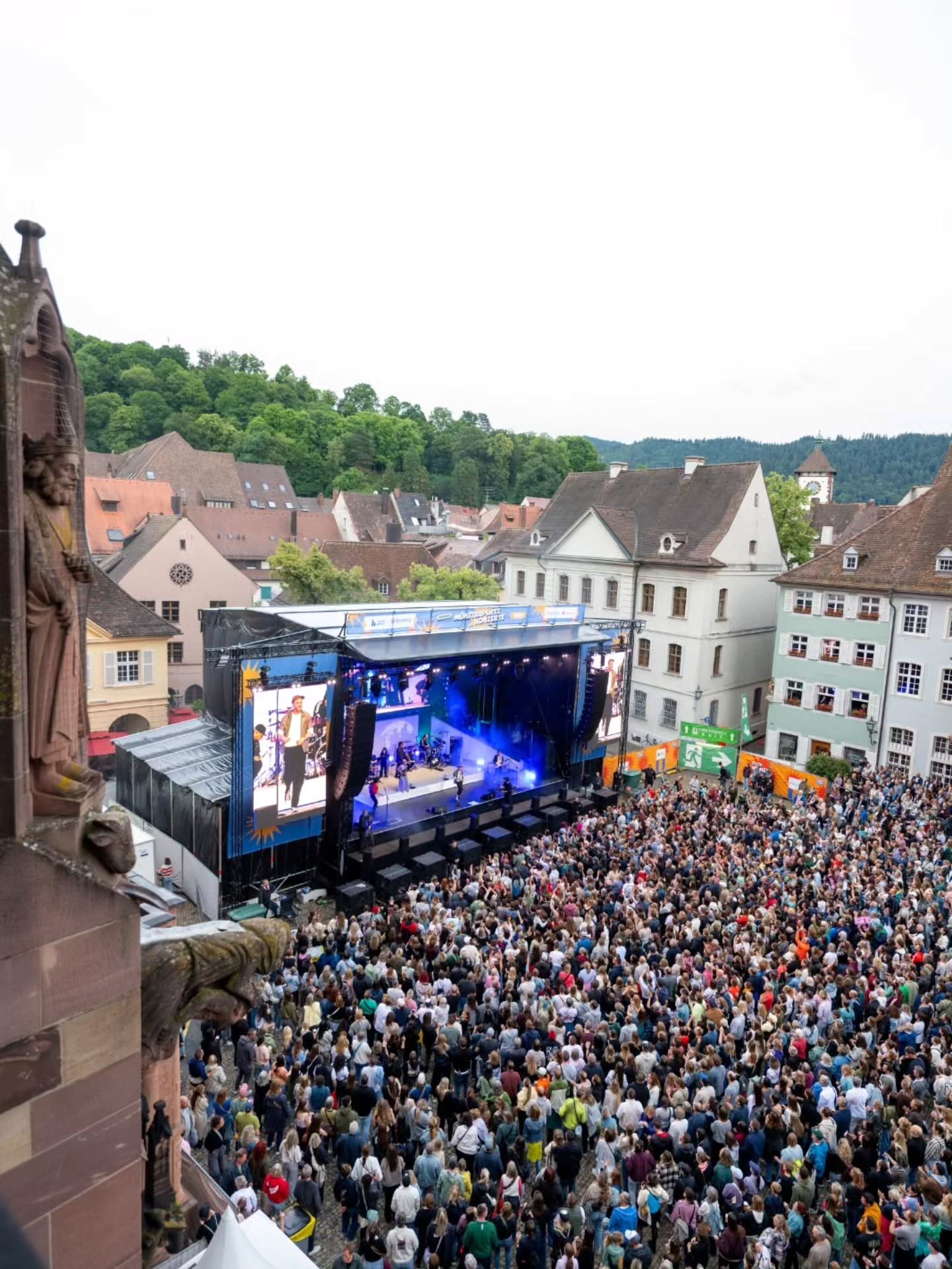 Große Menschenmenge vor einer Bühne mit Band und großen Bildschirmen in einer historischen Stadtmitte, im Hintergrund bewaldete Hügel. (KI-Alternativtext)
