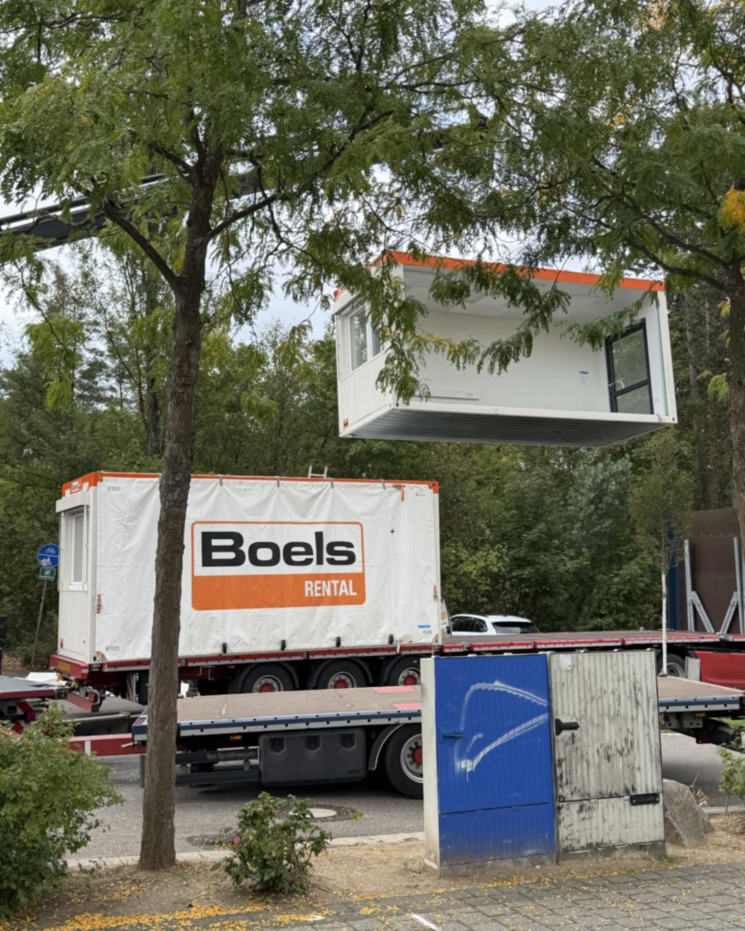 Ein weißer Container mit orangem Dachrand wird von einem Kran gehoben. Darunter steht ein LKW-Anhänger mit einem weiteren Container, auf dem groß "Boels RENTAL" steht. Im Vordergrund befindet sich ein blaues und weißes Tor vor Bäumen. (KI-Alternativtext)