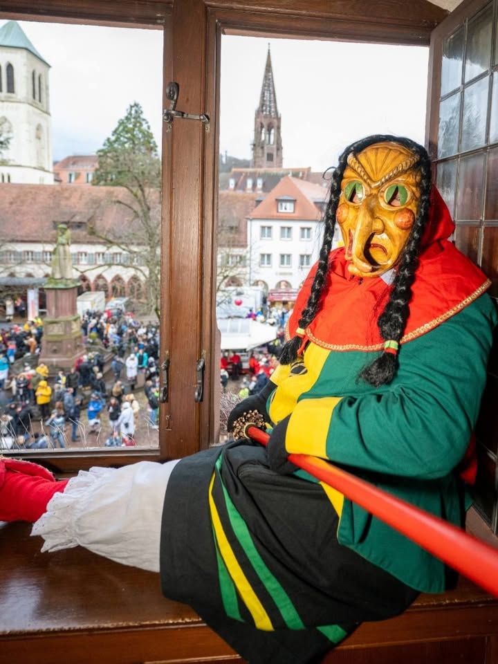 Dieser Alternativtext wurde KI-generiert: Eine Person mit einer verzierten Maske und einem farbenfrohen Kostüm sitzt in einem Fensterrahmen. Im Hintergrund ist eine Stadtansicht mit einer Kirche zu sehen.