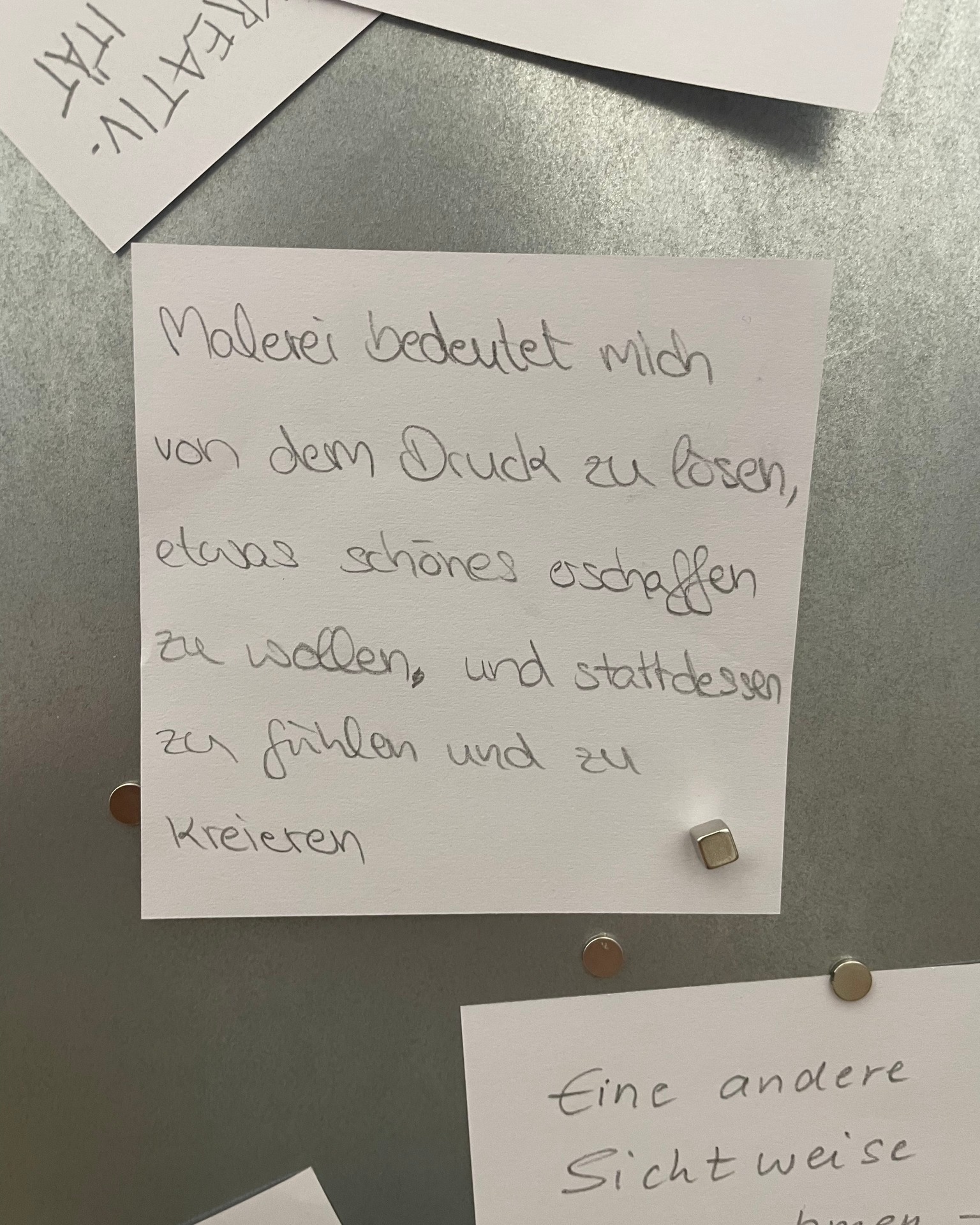 Dieser Alternativtext wurde KI-generiert: Ein gelbes Notizblatt mit handgeschriebenen Text in deutscher Sprache ist an einer grauen Oberfläche befestigt. Unterhalb befinden sich weitere, teilweise verdeckte Notizen.