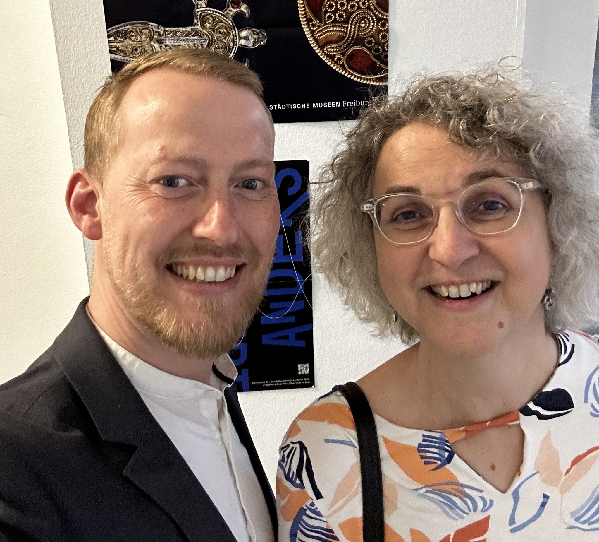 Dieser Alternativtext wurde KI-generiert: Zwei lächelnde Personen stehen nah beieinander. Im Hintergrund ist ein Museum zu sehen, mit einer Poster und einer kunstvollen Dekoration.
