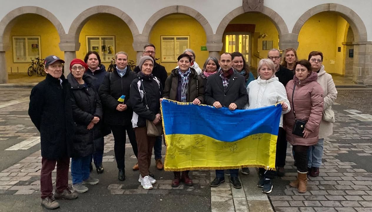 Dieser Alternativtext wurde KI-generiert: Eine Gruppe von Menschen steht vor einer gelben Fassade mit Arkaden zusammen. Sie halten eine blau-gelbe Fahne in den Händen.